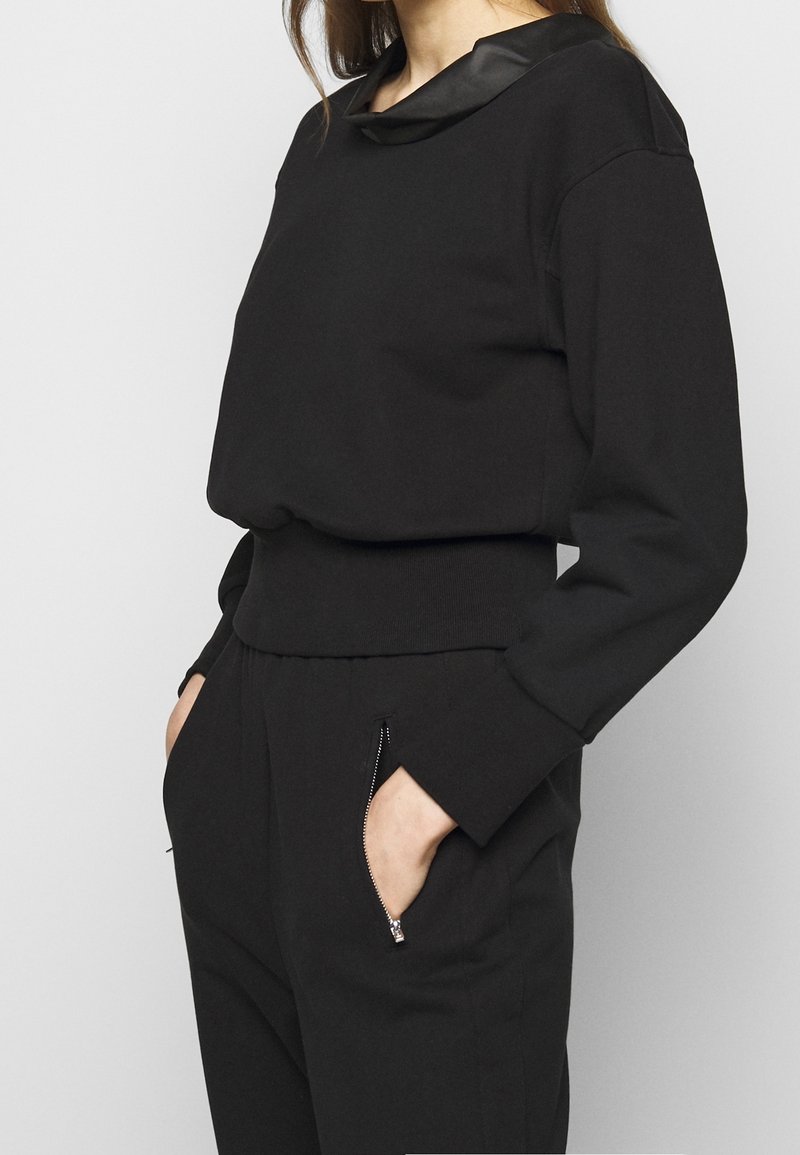 Černý top s dlouhým rukávem, se saténovým límečkem, žebrovaným pasem a kapsami. Hladká textura, volný střih a ploché švy.