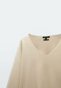 Maglione beige con scollo a V e maniche corte, realizzato in tessuto morbido. Design semplice e privo di decorazioni con una texture liscia. Etichetta visibile all'abbottonatura.