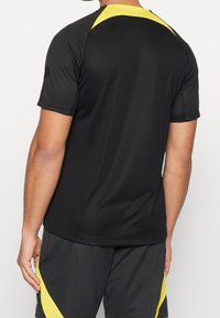 Camiseta deportiva masculina en negro con detalles amarillos, que presenta un patrón texturizado, mangas cortas y un diseño aerodinámico.