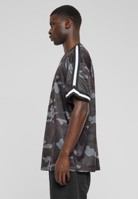 Camiseta de estampado camuflaje en tonos oscuros con acentos blancos en las mangas cortas. Hecha de tela de malla, con un ajuste relajado y holgado.