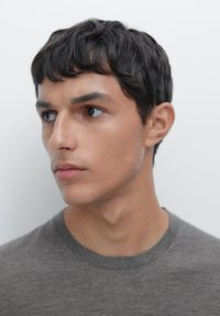 Ritratto di una persona con capelli corti e disordinati di colore scuro, che indossa una t-shirt grigia a girocollo. Messa a fuoco sui tratti del viso e sfondo neutro.