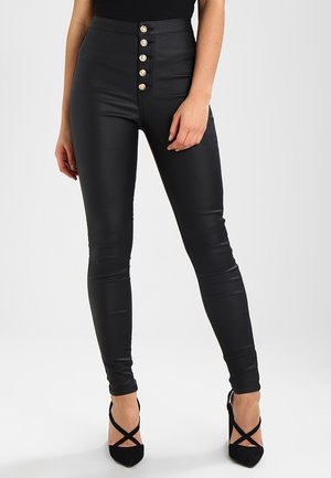 Woman wearing high-waist black leather skinny pants with five silver buttons and black pointed-toe heels with crisscross straps.