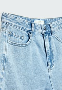 Jean en denim bleu clair montrant la partie avant de la taille avec un bouton, des passants de ceinture et une poche avant avec une petite poche à monnaie à l'intérieur.