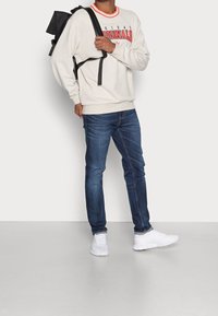 Sudadera crema con cuello rojo y estampado "Adidas Originals", jeans azules y zapatillas blancas. Mochila negra llevada sobre un hombro.