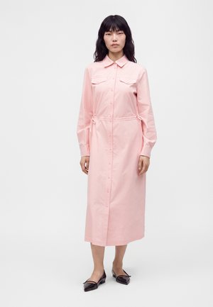 Vrouw met lang zwart haar, gekleed in een roze jurk met lange mouwen, knopen en borstzakken, en zwarte puntige platte schoenen, staand op een effen witte achtergrond.