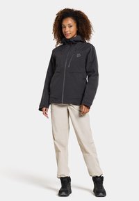 Veste imperméable noire avec capuche, poches zippées, poignets ajustables, portée sur un pantalon beige clair et des bottes de randonnée noires.