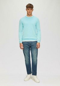 Hellblauer Sweatshirt mit langen Ärmeln und gerippten Bündchen, kombiniert mit ausgewaschenen blauen Jeans und weißen Turnschuhen, vor einem neutralen Hintergrund stehend.