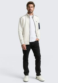 Witte ritsjacket met een gequiltde textuur en marineblauwe accenten, gedragen over een wit T-shirt, gecombineerd met zwarte jeans en witte sneakers.