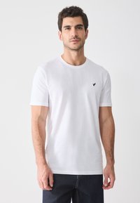 Weiße Baumwoll-T-Shirt mit kurzen Ärmeln, rundem Halsauschnitt und einem kleinen schwarzen Logo auf der linken Brust. Getragen mit dunklen Jeans.