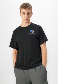 Joven con las manos en los bolsillos, vistiendo una camiseta negra de O'Neill con un gráfico de surf colorido y pantalones cargo grises, frente a un fondo blanco.