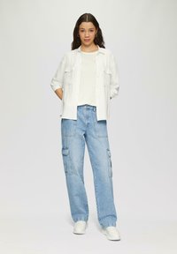 Witte katoenen overhemd met knopen, beige top en lichtblauwe cargobroek met zijzakken. Model draagt witte sneakers; effen lichte achtergrond.