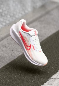 Deportivas blancas con parte superior de malla, acentos en rosa y logo de swoosh en rojo. Presentan una suela acolchada y diseño con cordones.