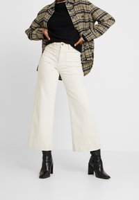 Vida vida byxa i krämfärg kombinerat med en stor rutig skjorta i brunt och gult, som kompletteras av svarta klackade ankelboots. Slät textur, avslappnad stil.