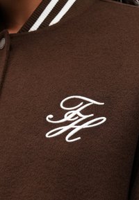 Chaqueta varsity marrón con letras entrelazadas bordadas en blanco "FH" en el pecho y cuello acanalado a rayas.