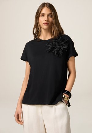 Schwarzes Baumwoll-T-Shirt mit kurzen Ärmeln, das mit einem strukturierten, schwarzen floralen Applikation auf der linken Schulter versehen ist. Kombiniert mit leichten, fließenden Hosen.