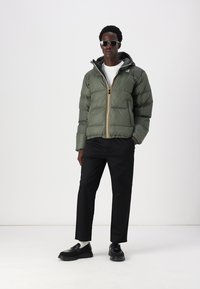 Giacca a vento verde oliva con zip gialla e nera, indossata sopra una camicia bianca. I pantaloni neri e le scarpe completano l'outfit.