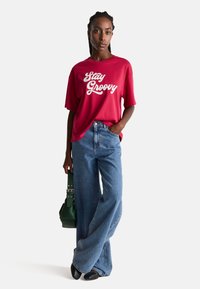 Czerwona, oversize'owa koszulka z napisem "Stay Groovy" w białym kolorze; w połączeniu z niebieskimi, wysokimi spodniami typu wide-leg i zieloną torebką. Czarne buty.