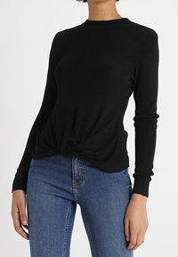 Femme portant un top noir à manches longues avec un détail de nœud sur le devant, associé à un jean en denim bleu, se tenant devant un fond blanc uni.