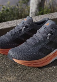 Paire de chaussures de course grises et orange reposant sur une surface en pierre à l'extérieur, avec une tige en maille texturée et des bandes latérales noires.