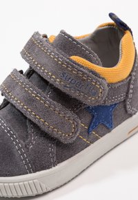 Scarpa per bambini in suede grigia con fodera gialla, dettaglio a stella blu, due cinturini in Velcro e suola in gomma bianca su sfondo bianco.