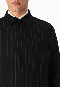 Juodas paltas su vertikaliomis rožinėmis kryptelėmis, turintis apykaklę, mygtukų uždarymą ir lygią tekstūrą. Išskirtinė detalė: logotipas šone.