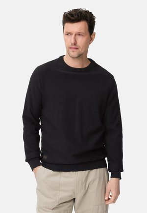 Man met kort krullend haar, gekleed in een zwarte trui zonder kraag en beige broek, staand met één hand in de zak, kijkt iets naar links.