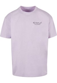 Lyslilla bomuld t-shirt med korte ærmer. Teksten på forsiden inkluderer arabisk skrift og den engelske sætning "Elsk dig selv først." Enkel design.