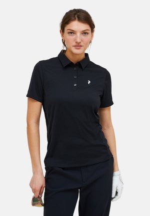 Schwarzes Poloshirt aus glattem Material, mit einem Kragen, drei Knöpfen und einem weißen Logo auf der linken Brust. Getragen mit dunklen Hosen und einem Handschuh.