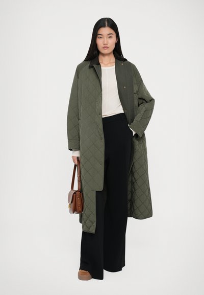Quiltet olivengrøn lang frakke med en mørk krave, båret over en lys top og sorte vide bukser. Accessories inkluderer en lille brun håndtaske.