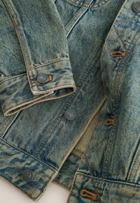 Közeli kép egy kifakult kék farmerdzseki ujjvégéről, amelyen patentos mandzsetták, fém patentgombok és narancssárga varrásrészletek láthatók.