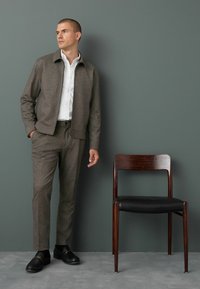 Chaqueta marrón texturizada con pantalones a juego, camisa blanca y zapatos negros. Silla de madera con asiento de cuero. Fondo neutro.