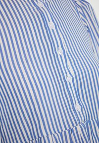 Blusa a righe in cotone bianco e blu. Presenta un colletto rotondo, chiusura con bottoni sul davanti e tessuto arricciato all'orlo.