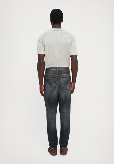 Uomo in piedi con le spalle alla macchina fotografica, indossa una maglietta a maniche corte grigia chiara, jeans scuri sbiaditi con tasche posteriori cucite e scarpe marroni.