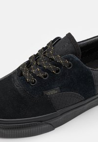 Baskets Vans noires avec panneaux texturés, lacets noirs et jaunes mouchetés, et semelle en caoutchouc sur fond blanc.