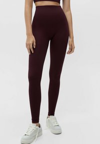 Leggings bordeaux taille haute en tissu extensible, avec une texture lisse et un design ajusté, associés à des baskets blanches.