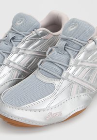 Ζευγάρι αθλητικά παπούτσια Asics σε ανοιχτό γκρι και ασημί με δίχτυ στη γλώσσα, κορδόνια και ανοιχτό καφέ σόλες, εμφανιζόμενα από πλάγια πάνω όψη.
