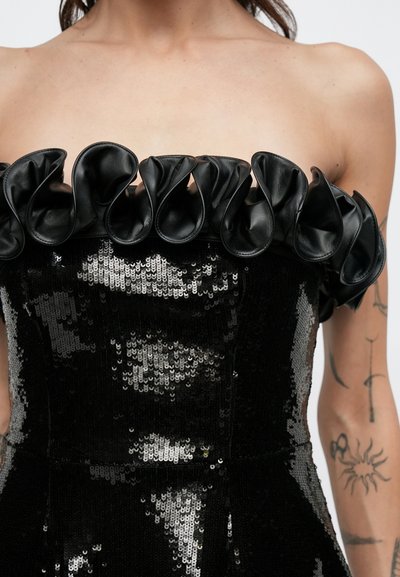 Melna bezplecu kleita ar spīdīgu aplikāciju faktūru, cieši pieguļošu korseti un ādas kroku akcentiem gar izgriezumu.