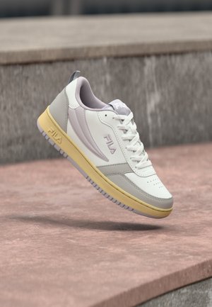 Biało-szary sneaker FILA z żółtą podeszwą unoszący się nad różową kamienną powierzchnią i szarym kamiennym tłem.