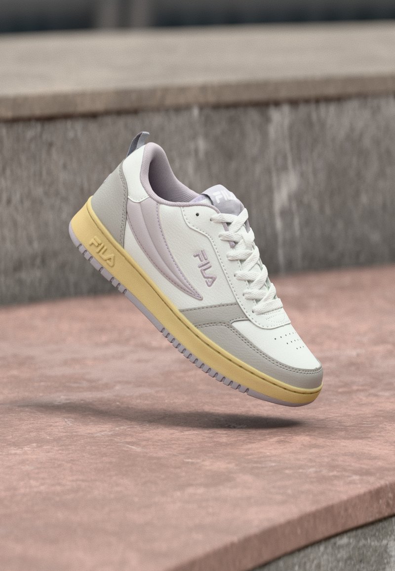 Λευκό και γκρι αθλητικό παπούτσι FILA με κίτρινη σόλα που αιωρείται πάνω από μια ροζ πέτρινη επιφάνεια και φόντο από γκρι πέτρες.