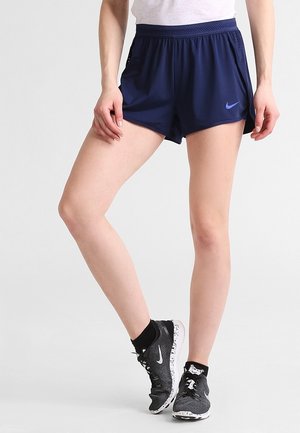 Nogi w granatowych sportowych szortach Nike, czarnych skarpetkach za kostkę oraz czarno-białych butach do biegania Nike na jednolitym tle.