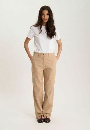 Femme debout, les mains dans les poches, portant un t-shirt blanc, un pantalon beige et des chaussures plates tissées foncées sur un fond clair uni.