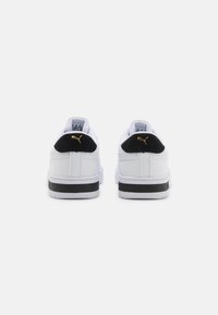 Ett par vita sneakers med svarta hälflikar som har guldiga Puma-logotyper, sedda bakifrån på en vit bakgrund.