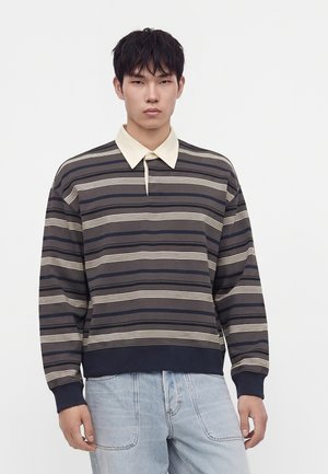 Jeune homme portant un maillot de rugby rayé à manches longues avec un col beige et un jean bleu clair sur fond blanc.