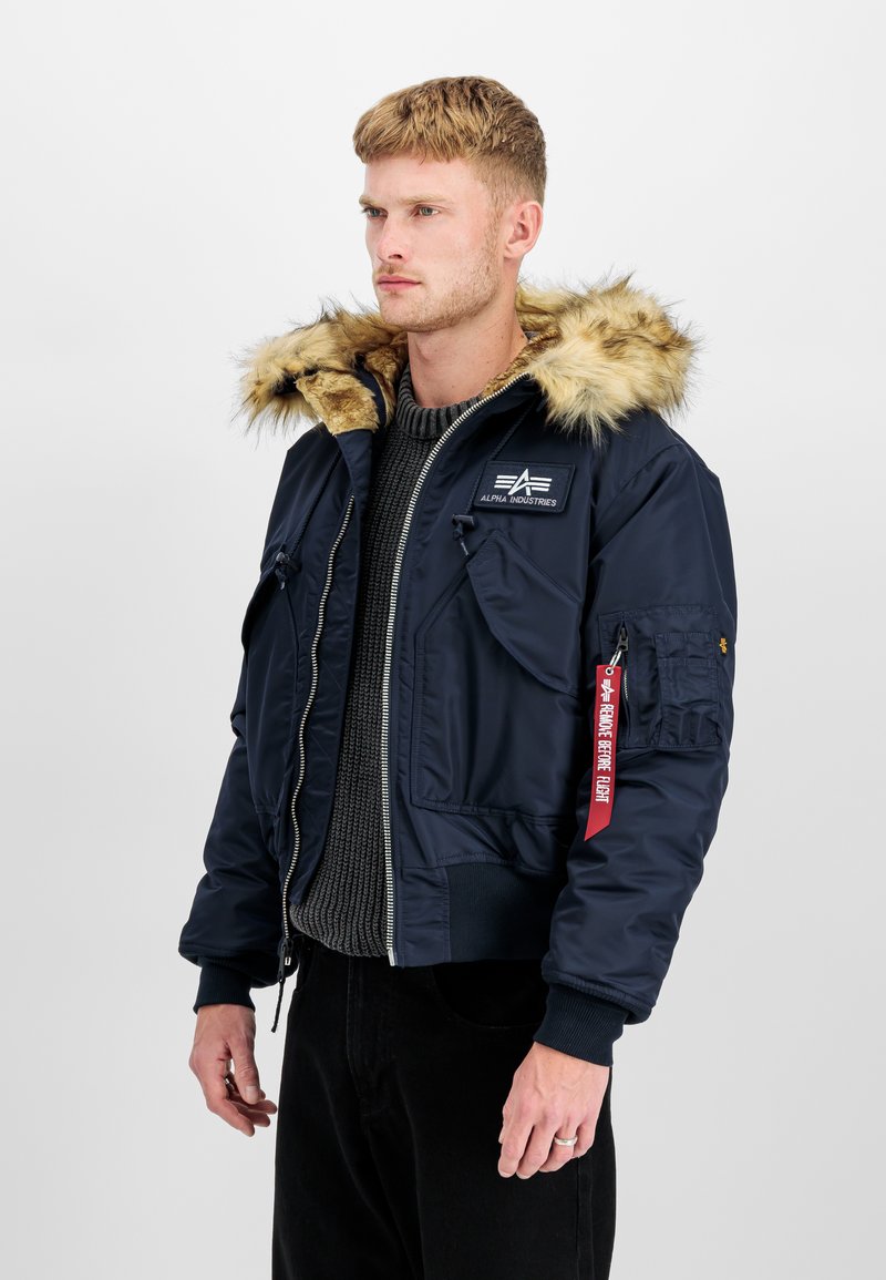 Junger Mann trägt eine marineblaue Alpha Industries Jacke mit Kapuze aus Fell, grauen Pullover und schwarze Hose, steht vor einem einfarbigen Hintergrund.