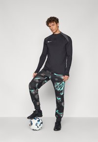 Sort langærmet træningsskjorte med en halv lynlås og grå skulderdetaljer, parret med mønstrede sorte sweatpants med teal design.