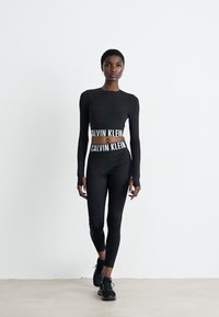 Eine Frau läuft nach vorne und trägt ein schwarzes langärmeliges Cropped-Top von Calvin Klein sowie hochgeschnittene Leggings und schwarze Sneakers vor einem weißen Hintergrund.