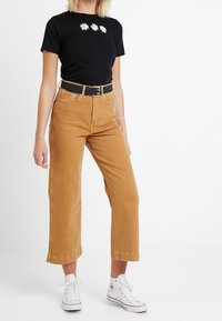 Camiseta negra corta con tres diseños de margaritas blancas, combinada con unos jeans de pierna ancha color tostado de cintura alta y zapatillas blancas. Cinturón negro como detalle.