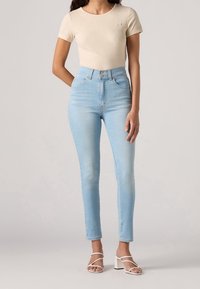 Vaqueros de tiro alto azul claro con ajuste slim, con cierre de botones y un sutil logo de la marca en una camiseta beige ajustada.