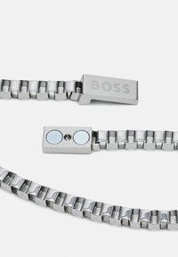 Bracelet en métal argenté avec des maillons entrelacés, doté d'un fermoir rectangulaire gravé de "BOSS" et d'une fermeture magnétique avec deux accents ronds.