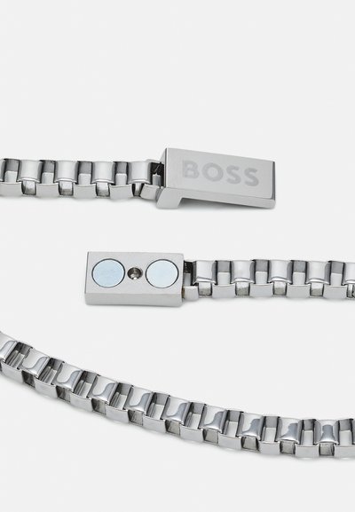 Bracelet en métal argenté avec des maillons entrelacés, doté d'un fermoir rectangulaire gravé de "BOSS" et d'une fermeture magnétique avec deux accents ronds.